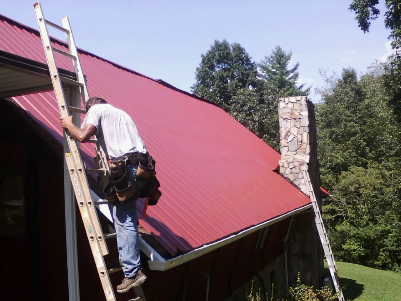 Roofer on a ladder beside a red metal roof and stone chimney — Soffit & Fascia Repair by Roofing Brothers in Woodford